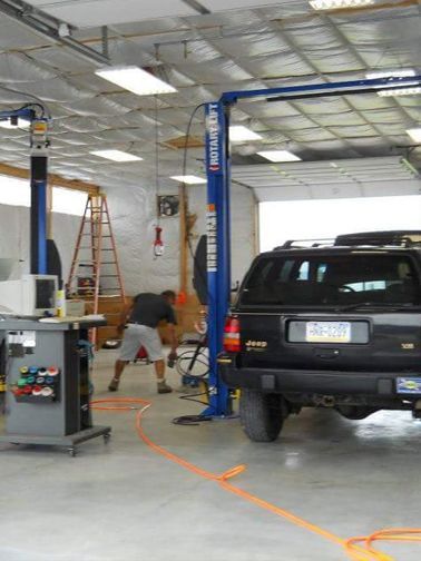 A mechanic working on a vehicle inside a workshop. Car is on a lift, worker in shorts. Orange hose on the floor.