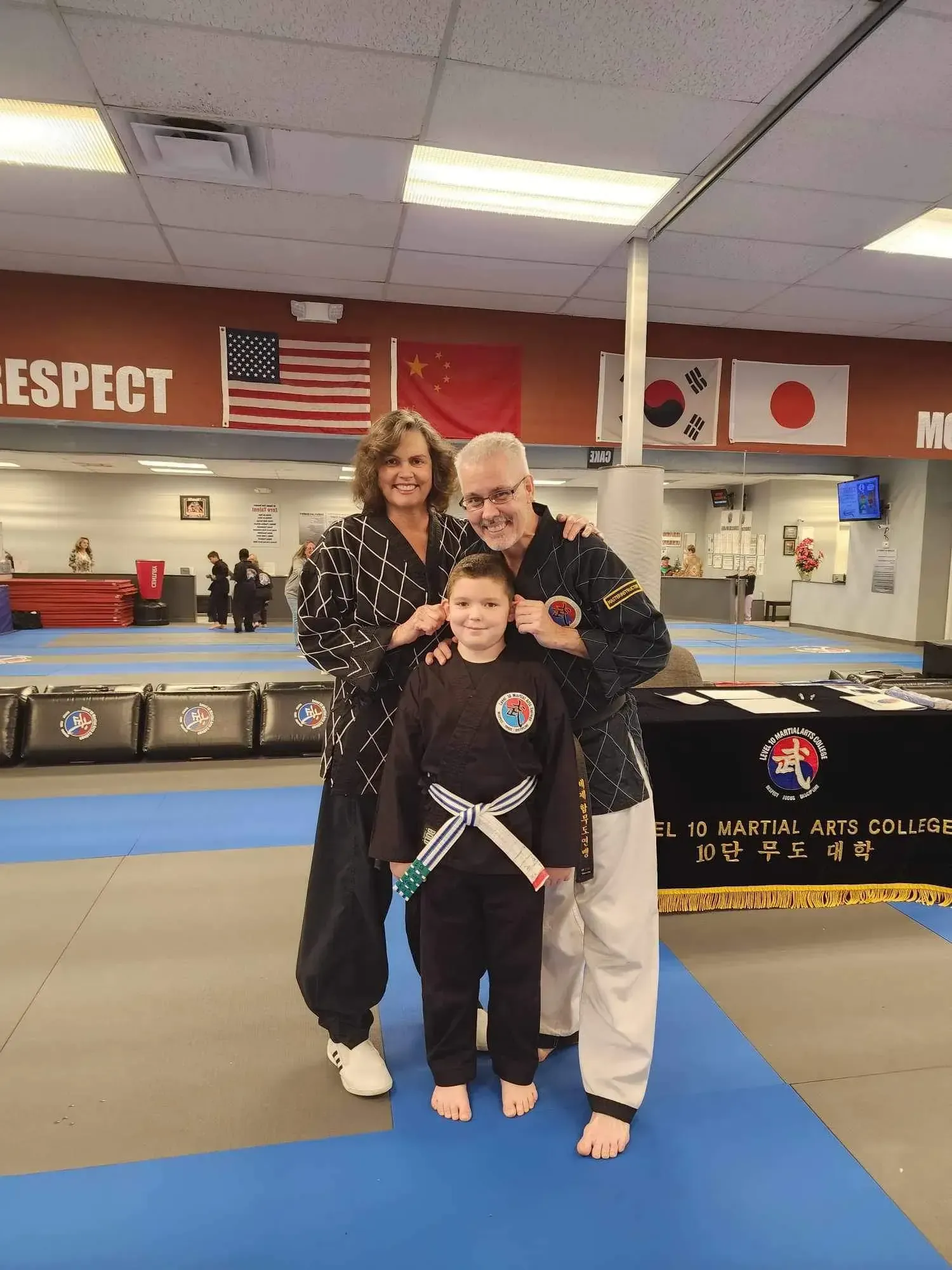 Three people in martial arts uniforms pose in a dojo with blue mats and flags on the wall.