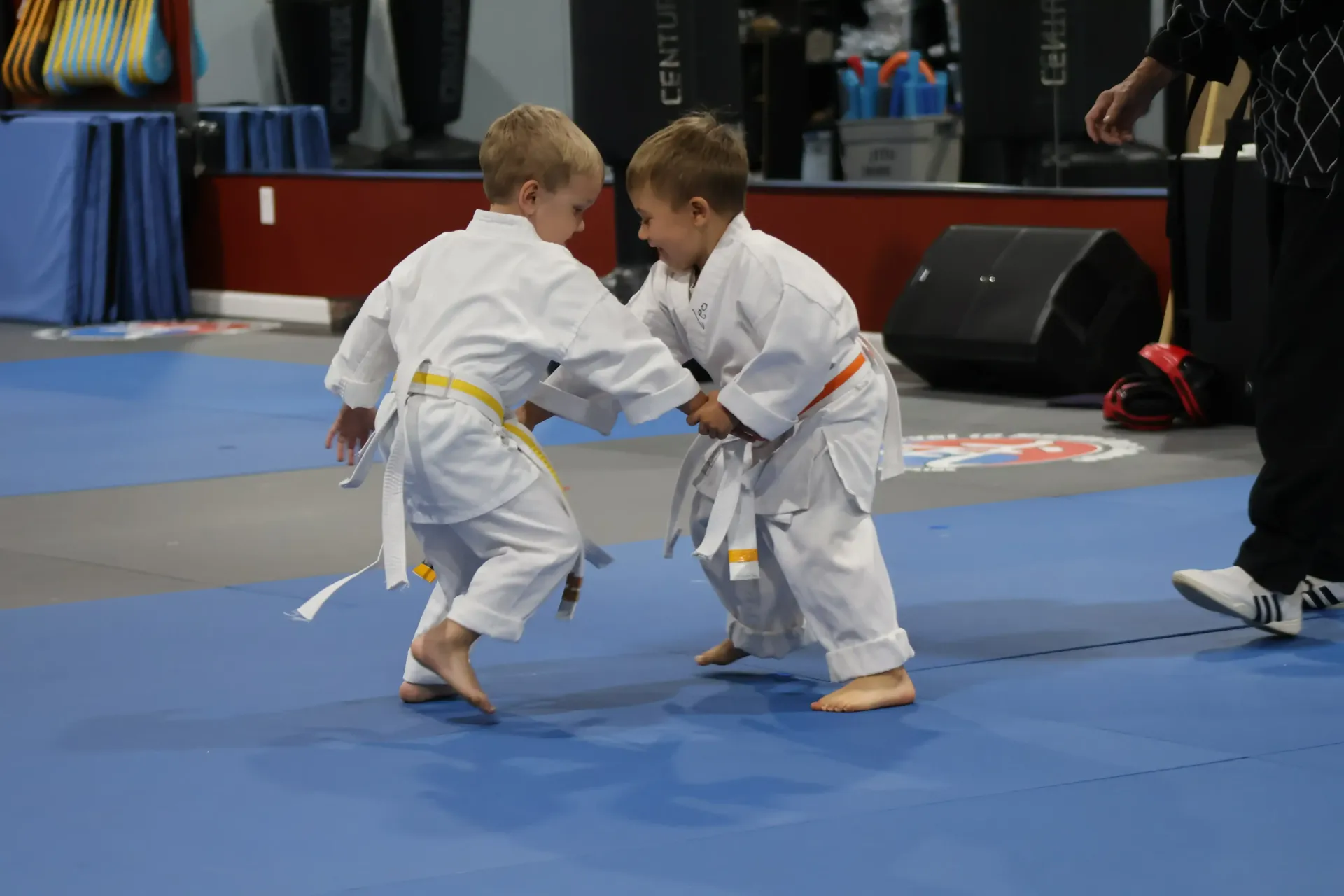 Two children in white martial arts uniforms sparring on a blue mat at an indoor competition