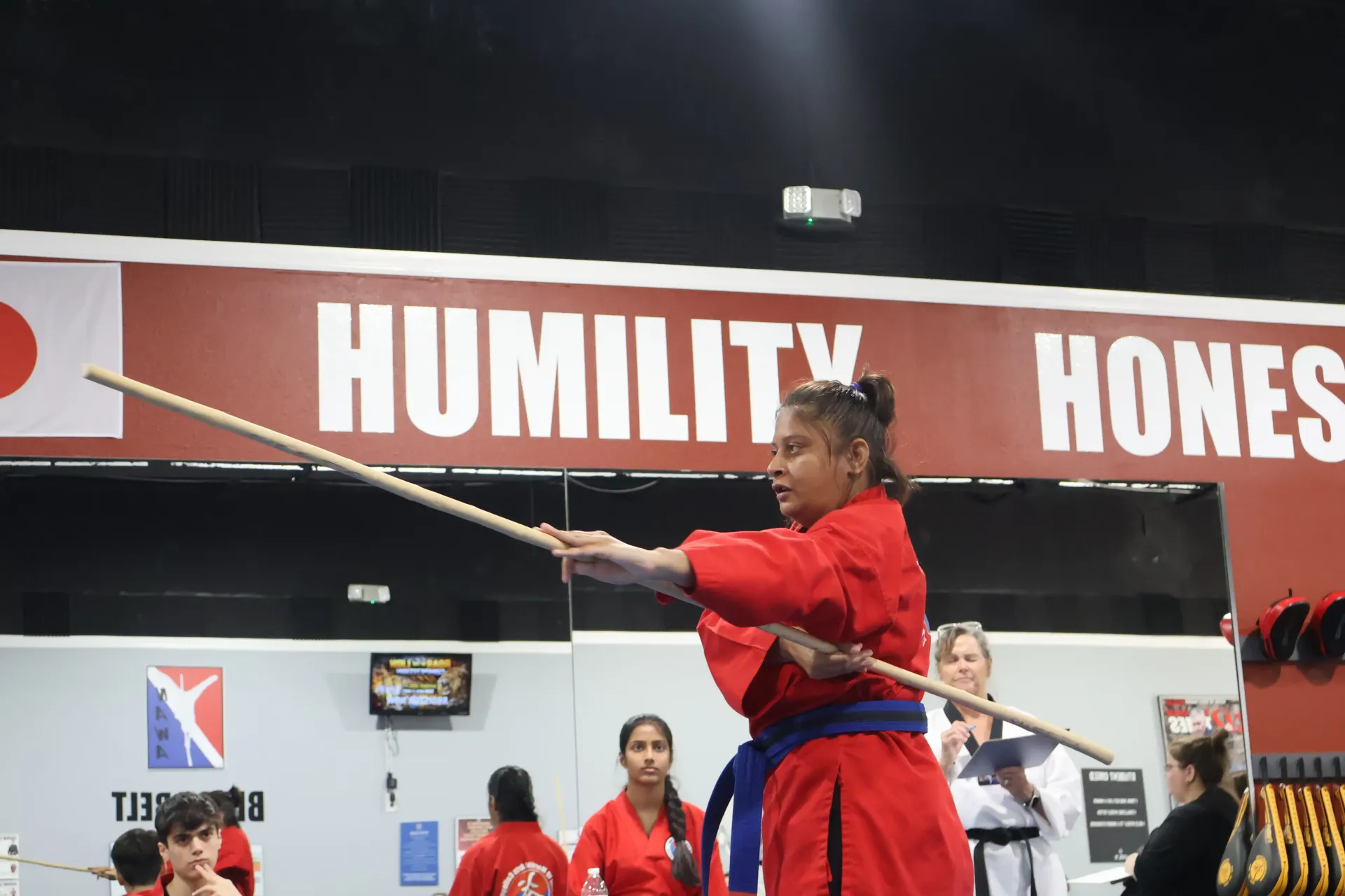 Martial artist in a red uniform practicing staff form indoors under a “HUMILITY” banner