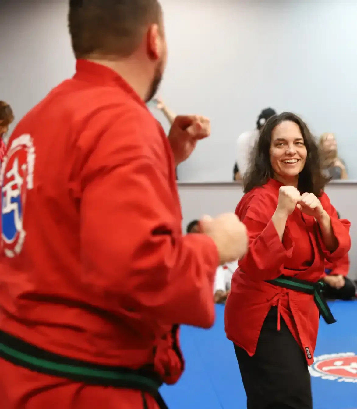 Two people in red martial arts uniforms spar indoors, smiling with raised fists.