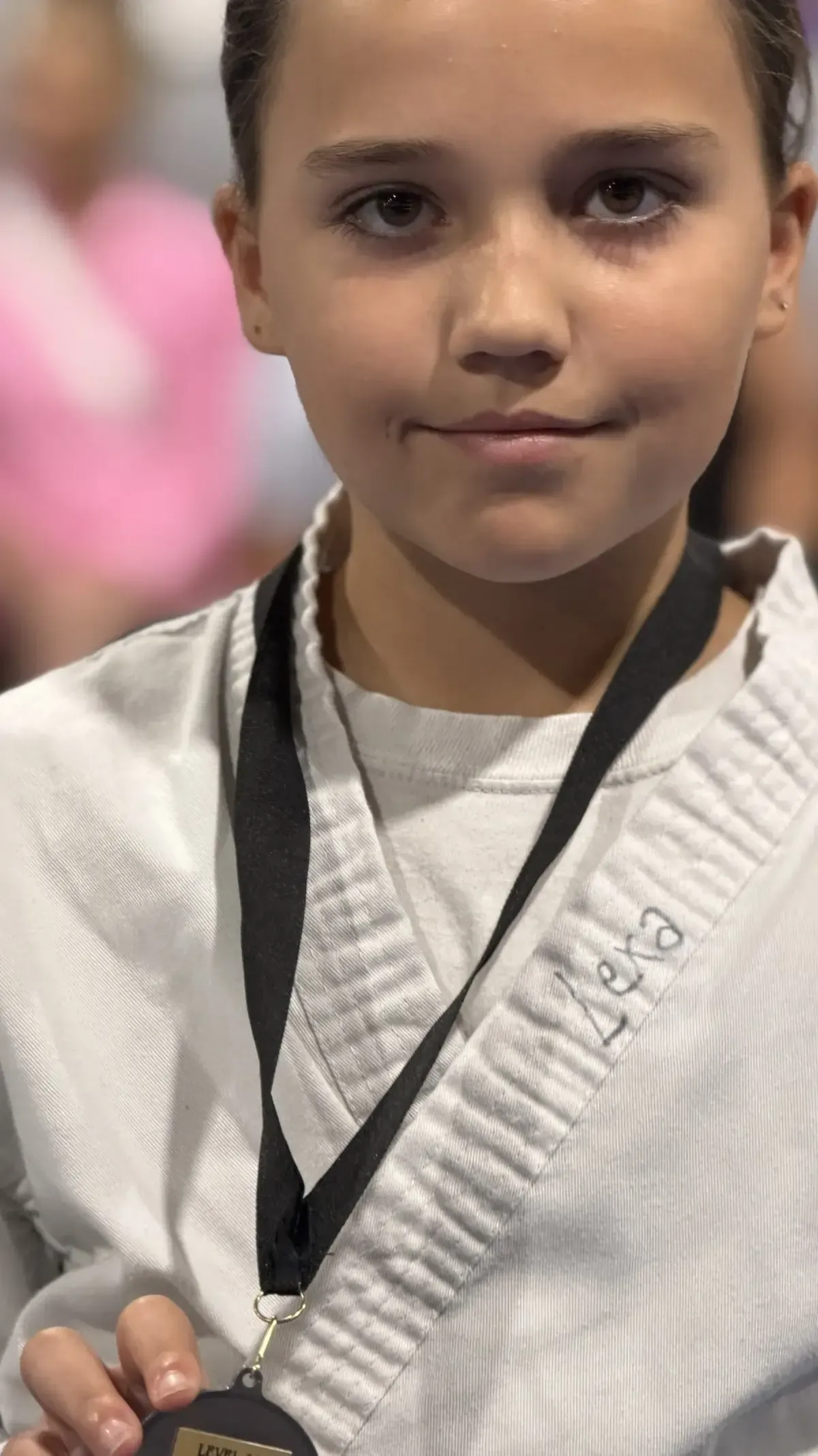 Young martial artist in a white gi with black belt, facing camera and holding a medal by its ribbon.