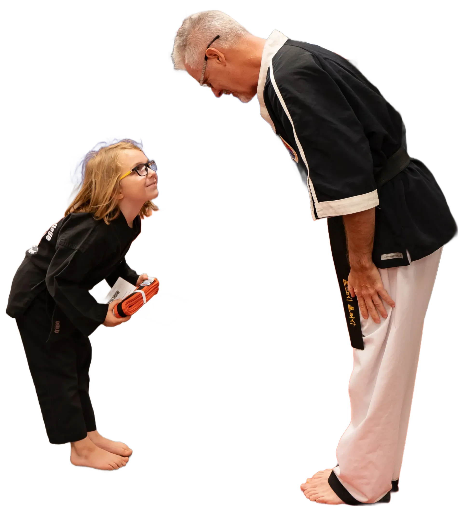 Two people in black uniforms face each other, one bowing while holding an orange object in a plain white studio.