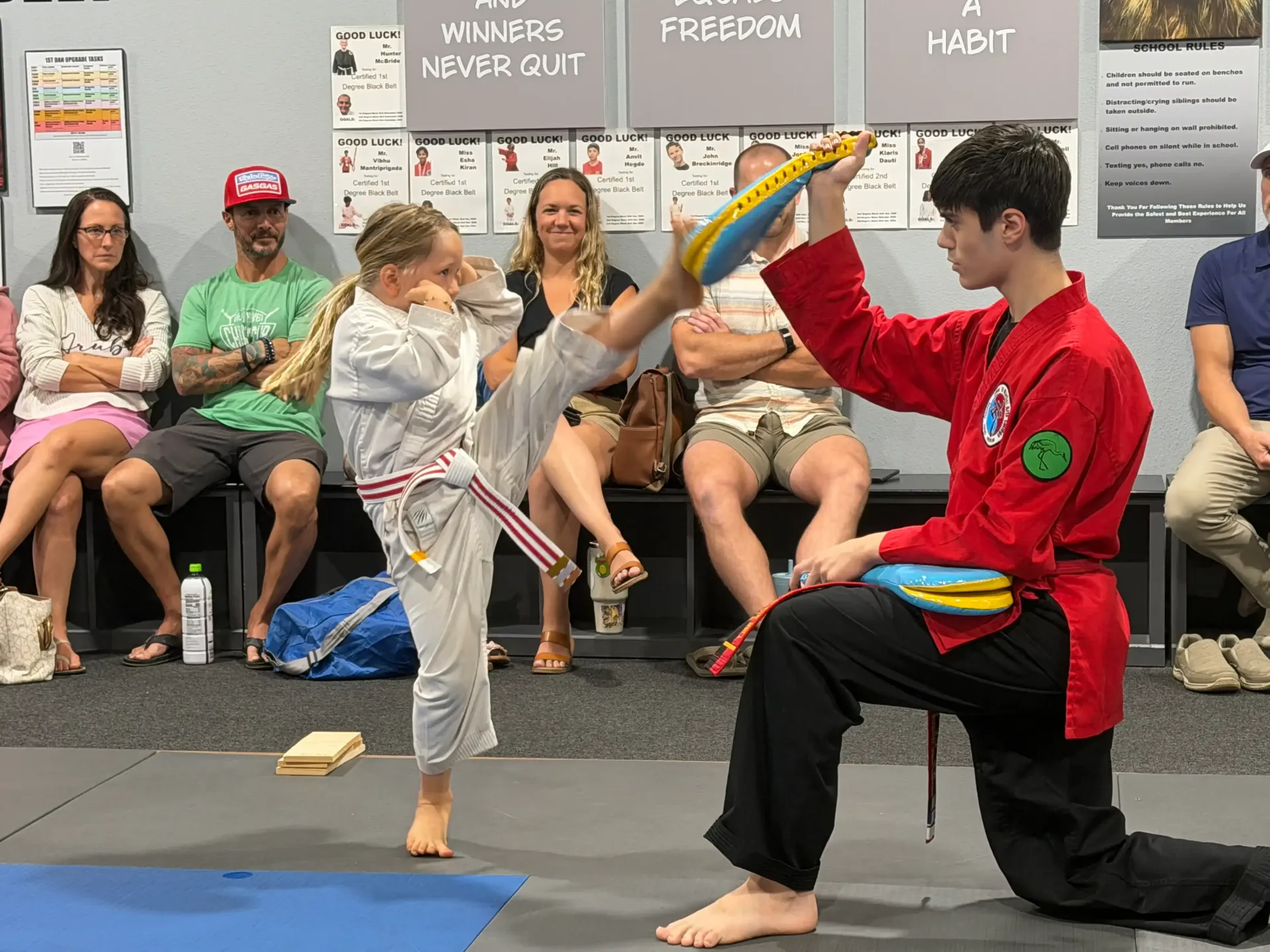 Two people practicing a martial arts move in a room while seated spectators watch behind them.