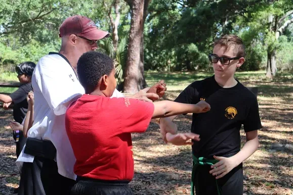 Three people chatting outdoors in a park, one in a red shirt gesturing toward a man in a black shirt.