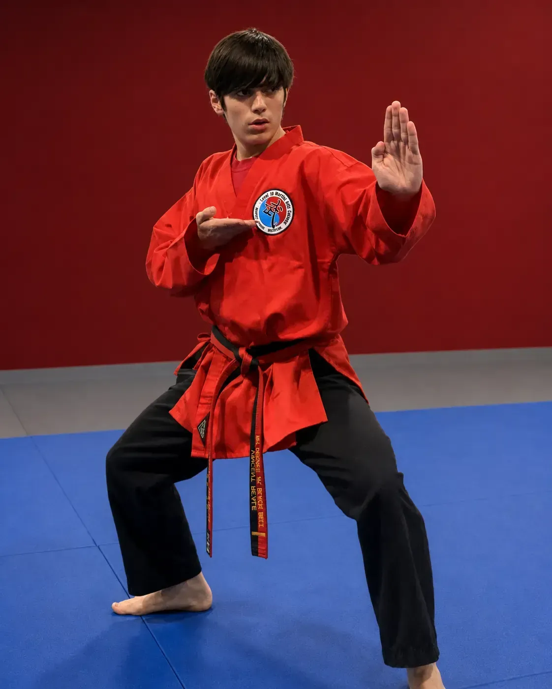 Martial artist in red uniform holding a Taekwondo block on blue mat against a red backdrop