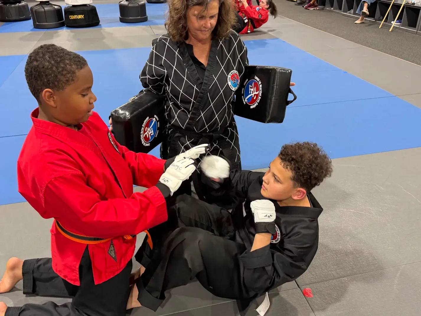 Children practicing martial arts with a coach in a gym, one holding pads while another strikes.