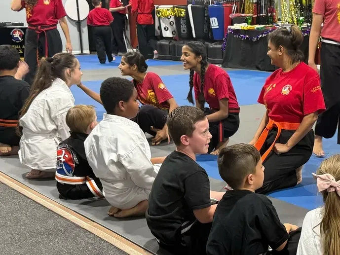 Children seated on mats watching two women in red shirts lead an activity in a gymnasium