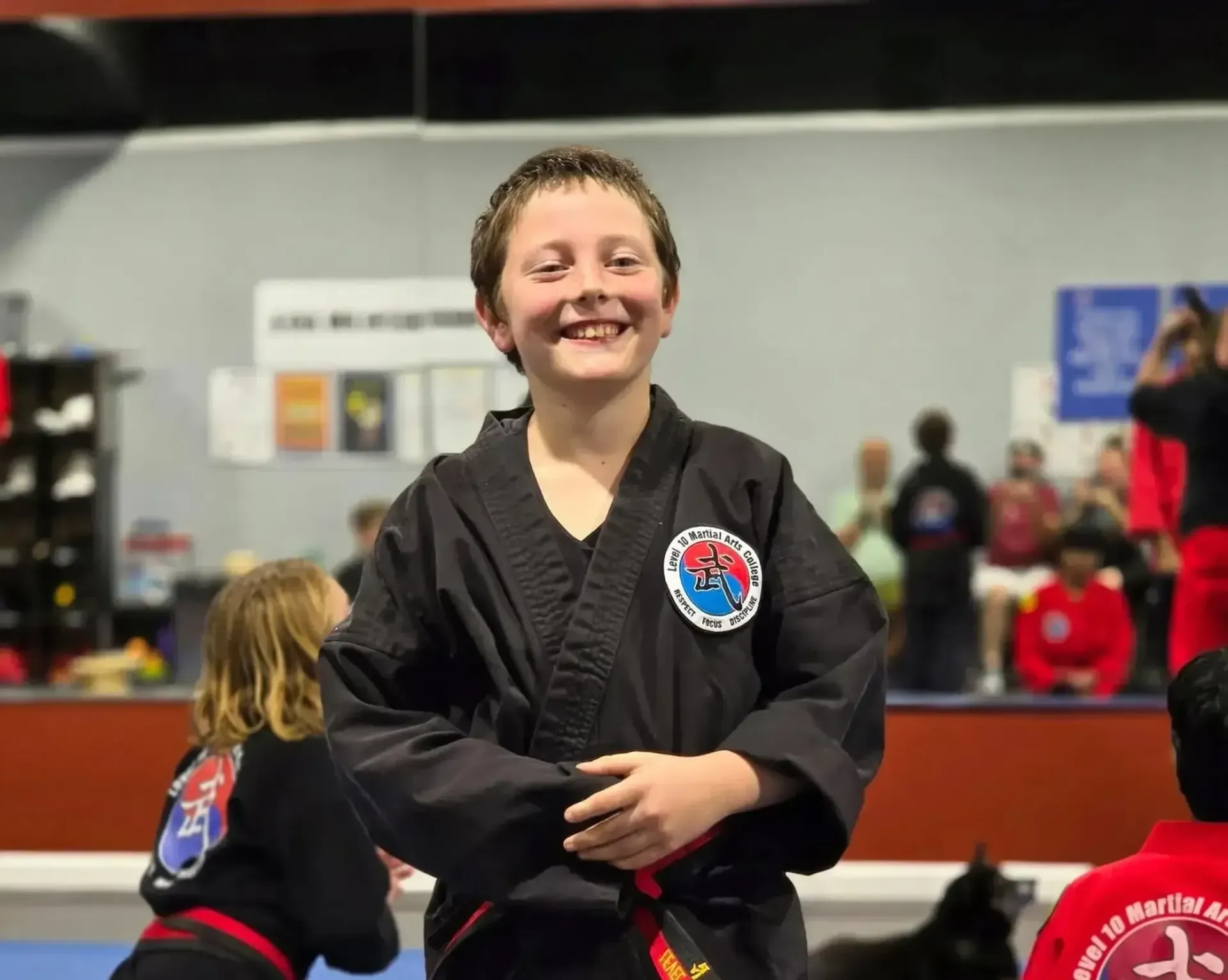 Smiling martial arts student in a black uniform standing in a dojo, with others blurred in the background