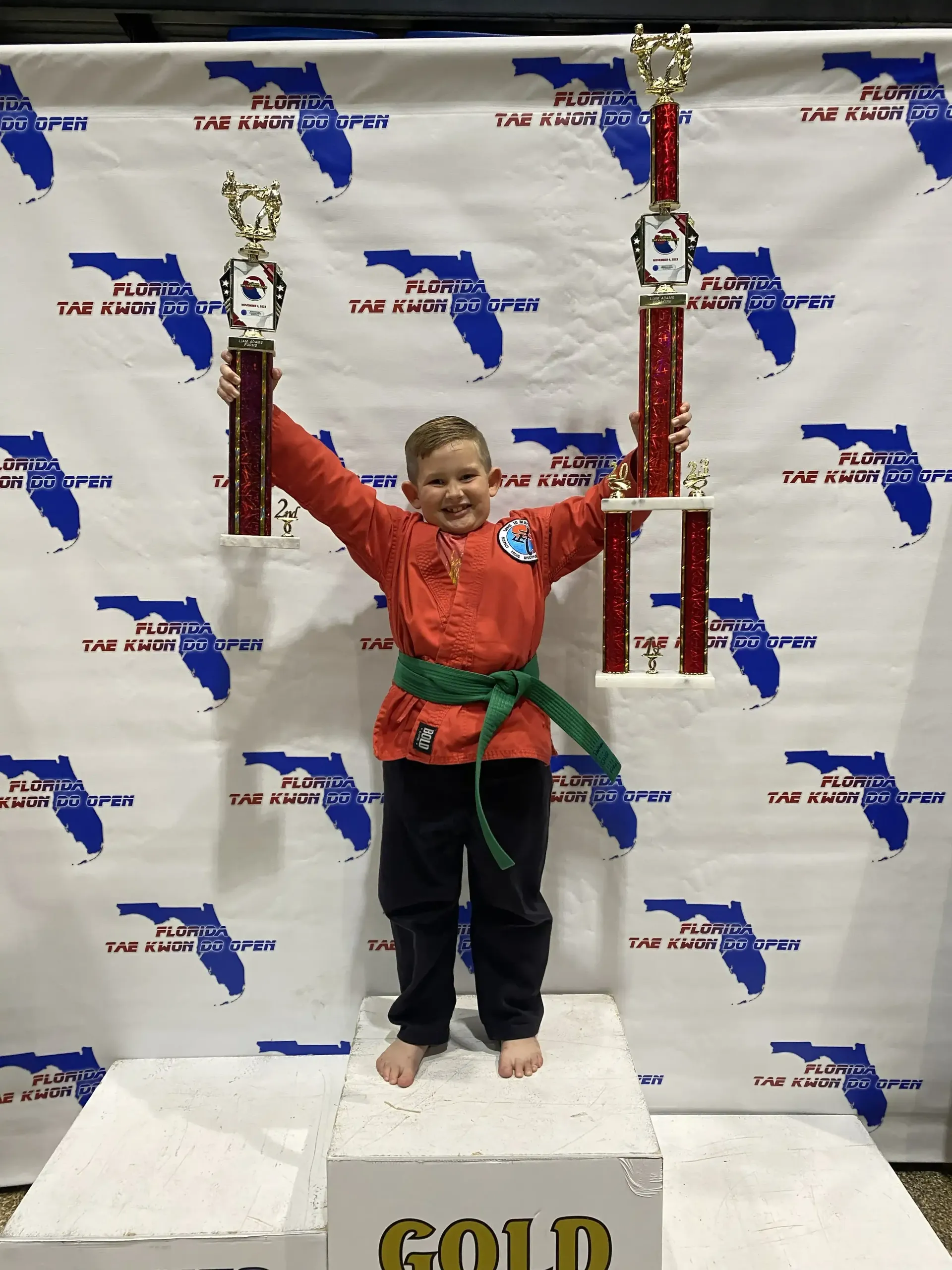 Child in red and green martial arts uniform holding trophies on a winners podium with a gold
