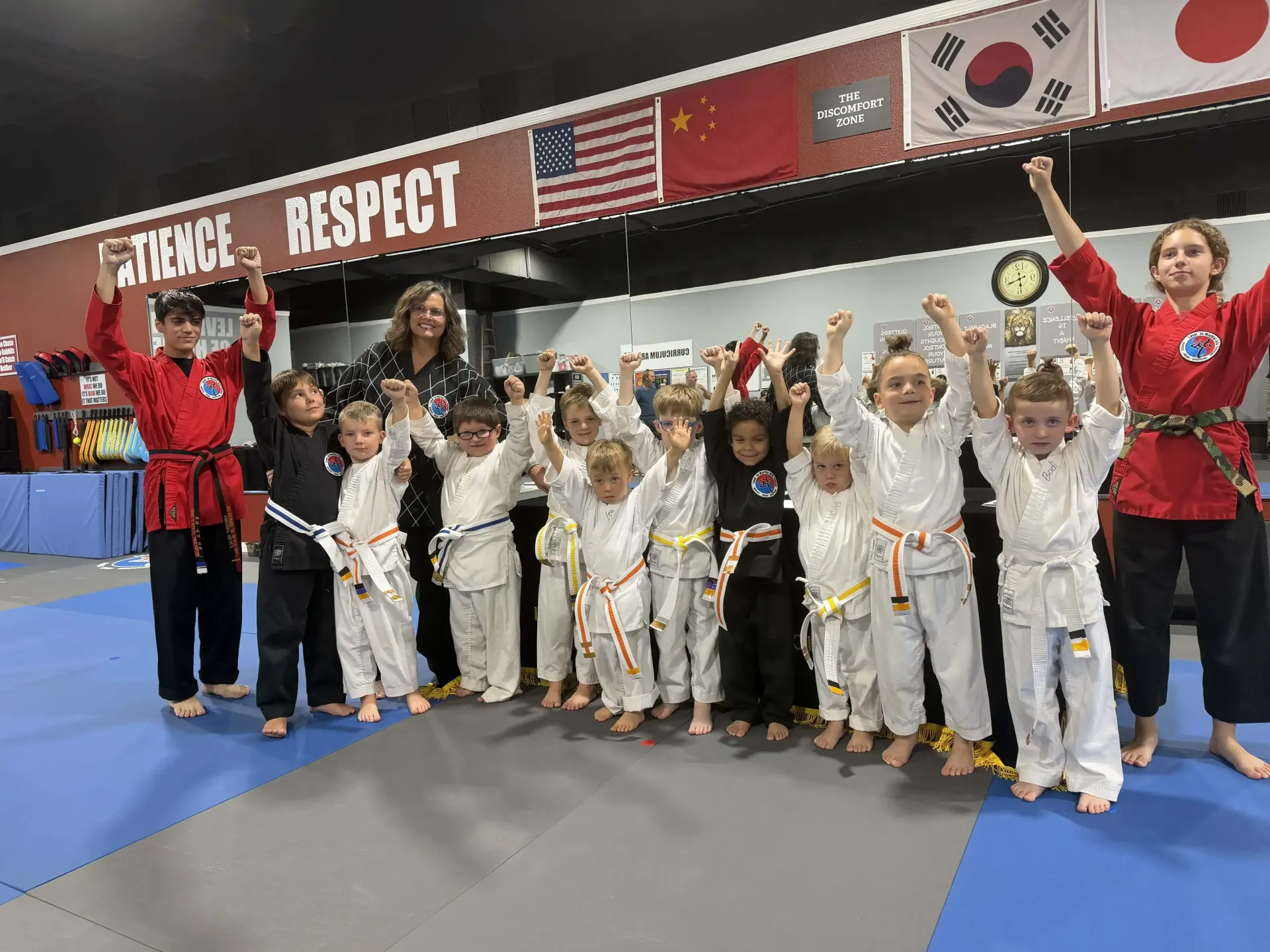 Children and instructors in white martial arts uniforms pose in a gym under “Respect” and flag banners.