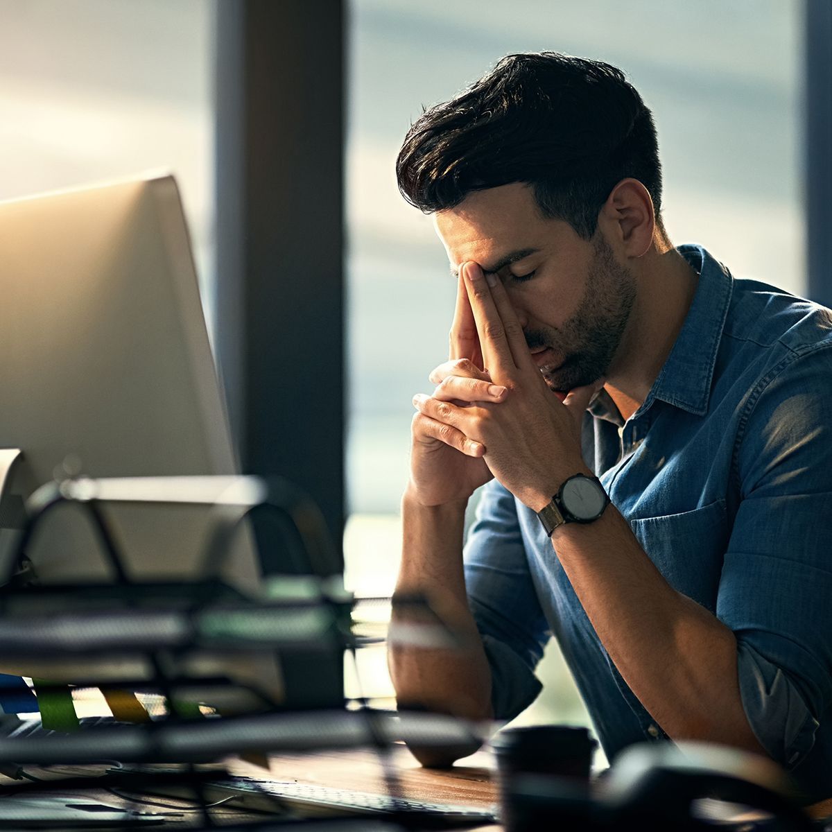 Man in blue shirt sits at desk, head bowed and hands clasped, looking stressed in an office.