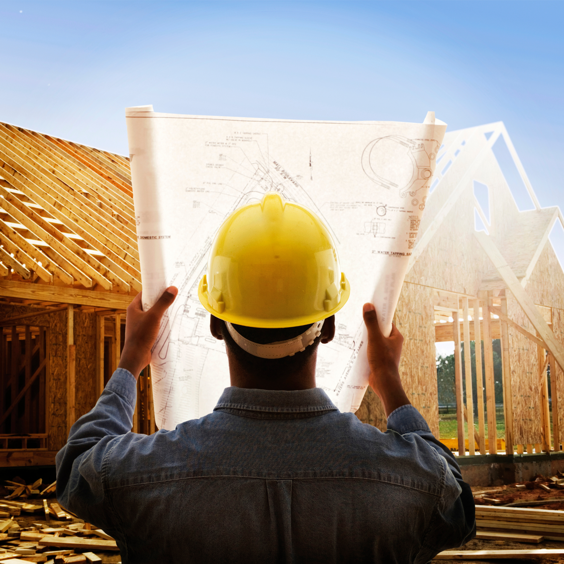 A man wearing a hard hat is holding a blueprint in front of a house under construction