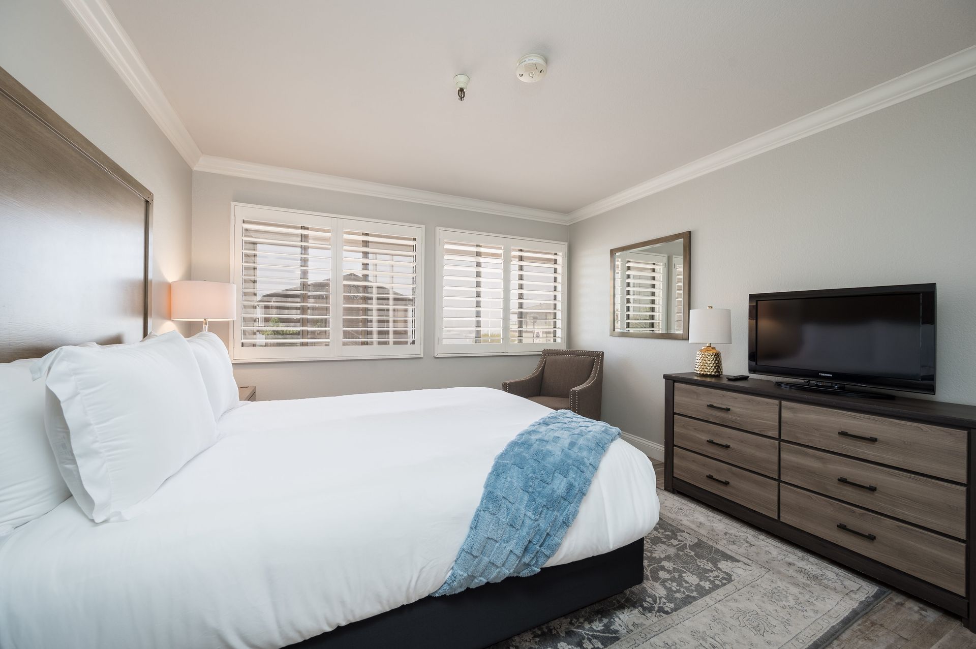 A hotel room with a king size bed , dresser , chair and television.