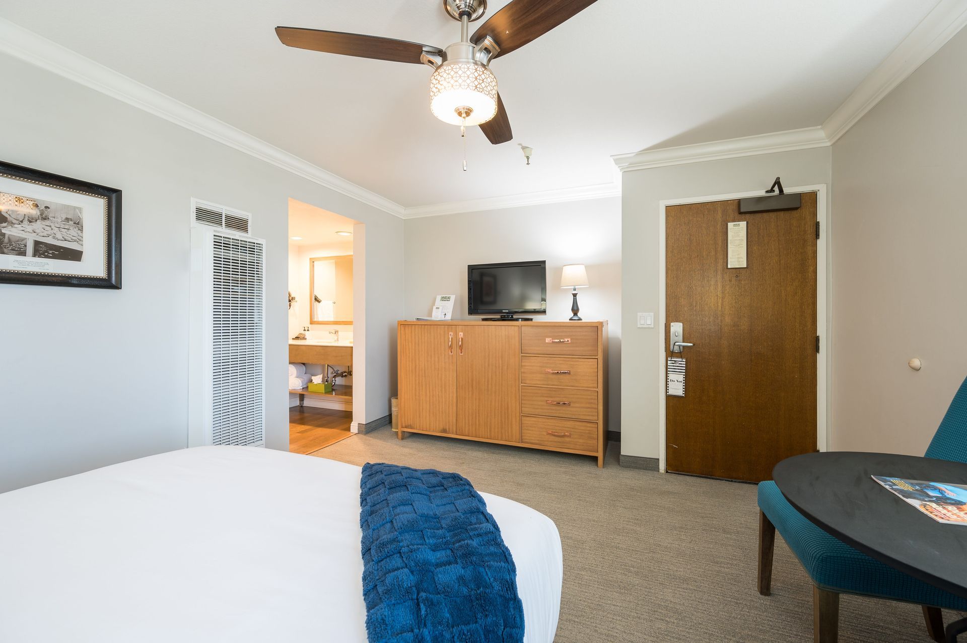 A hotel room with a king size bed , ceiling fan , television and dresser.