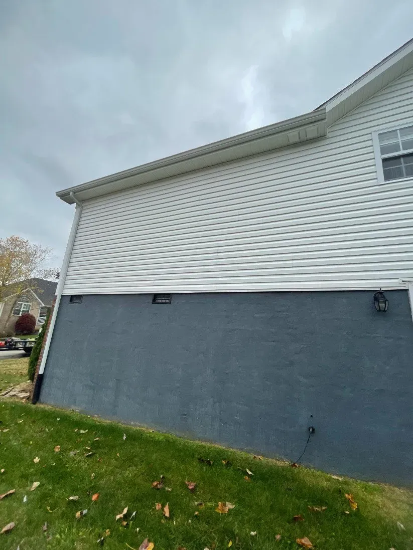 A white house with a gray siding is sitting on top of a lush green hillside.
