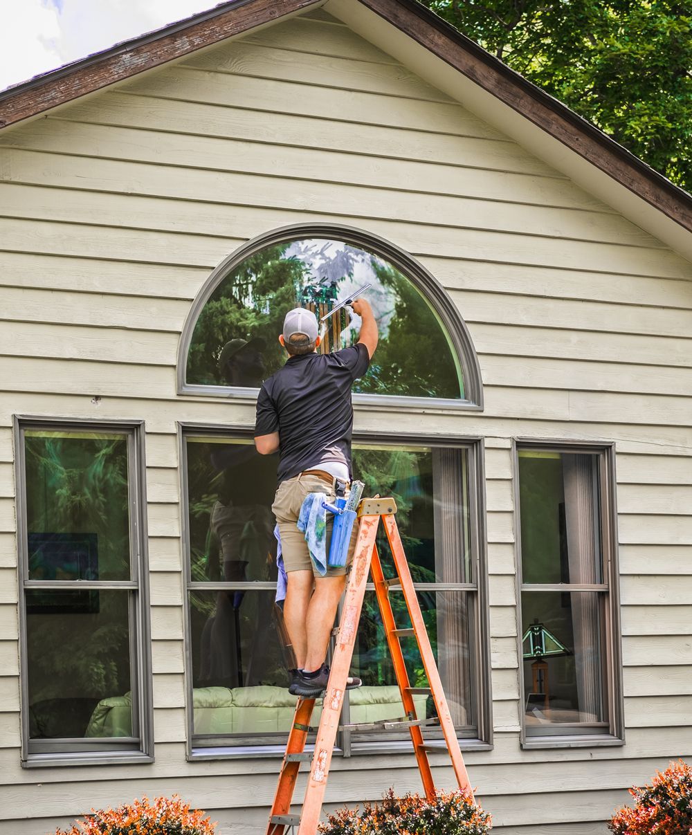 A man is standing on a ladder cleaning a window on the side of a house.