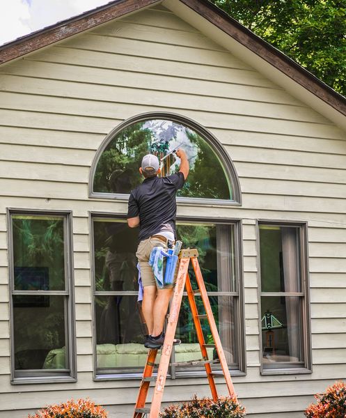 A person cleans a large arched window from a ladder on a house's exterior.