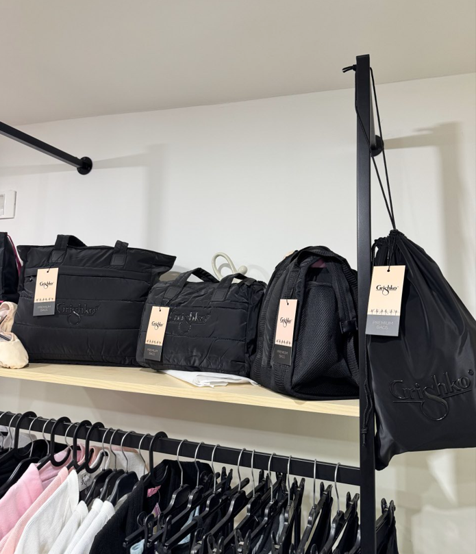 Borse nere su uno scaffale di un negozio, tra cui una borsa tote, una borsa da viaggio e una borsa con coulisse; sotto, vestiti appesi alle grucce.