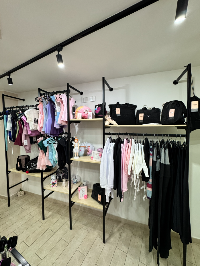 Interno di un negozio di abbigliamento, espositori con indumenti, scaffali di colore chiaro, struttura in metallo nero, pareti bianche, illuminazione dall'alto.