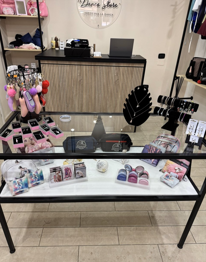 Un tavolo espositivo con vari prodotti, tra cui gioielli e piccole borse. Dietro l'espositore si trovano un registratore di cassa e un computer portatile.