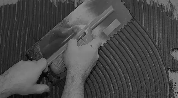 Eine Person trägt mit einer Kelle Klebstoff auf einen Fliesenboden auf.