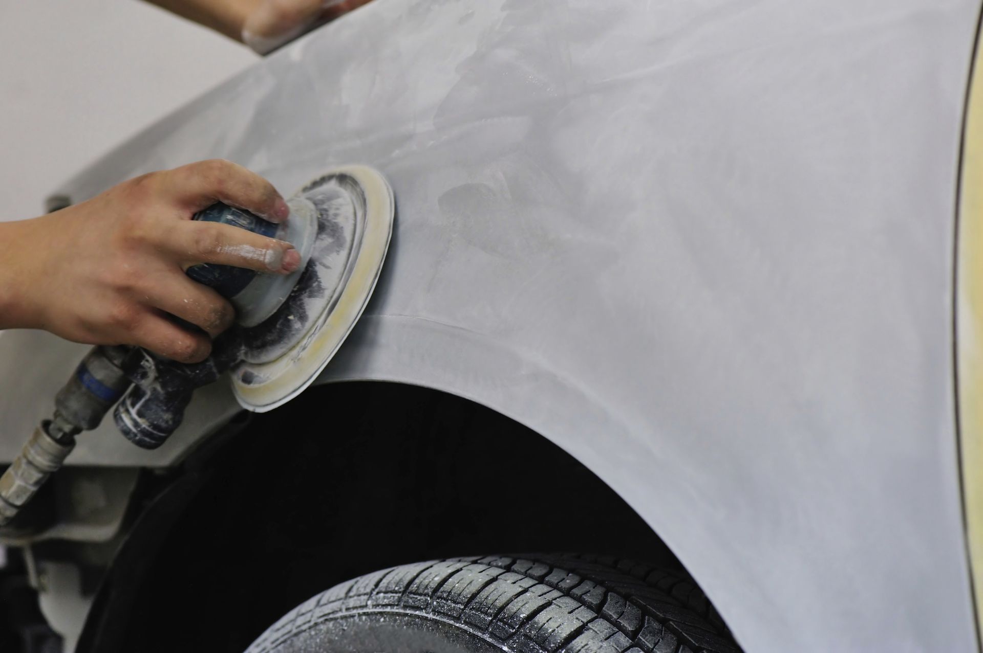 A person using a sander on a car fender, bodywork in progress, near a tire.