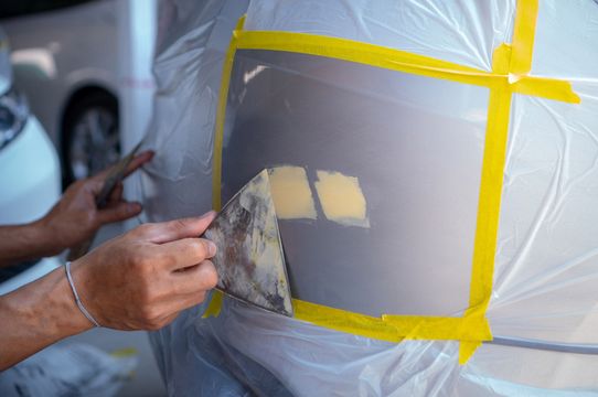 Person using a putty knife on car panel; masked for paint, two filled areas visible.
