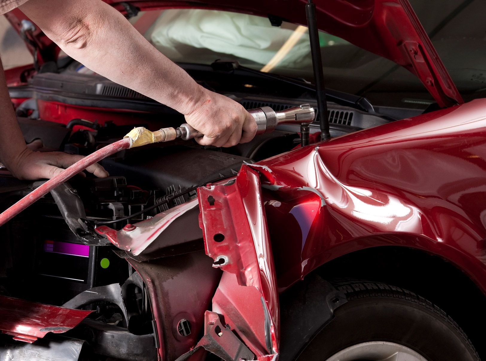 Mechanic repairs a damaged red car in a garage using a tool connected to a hose.