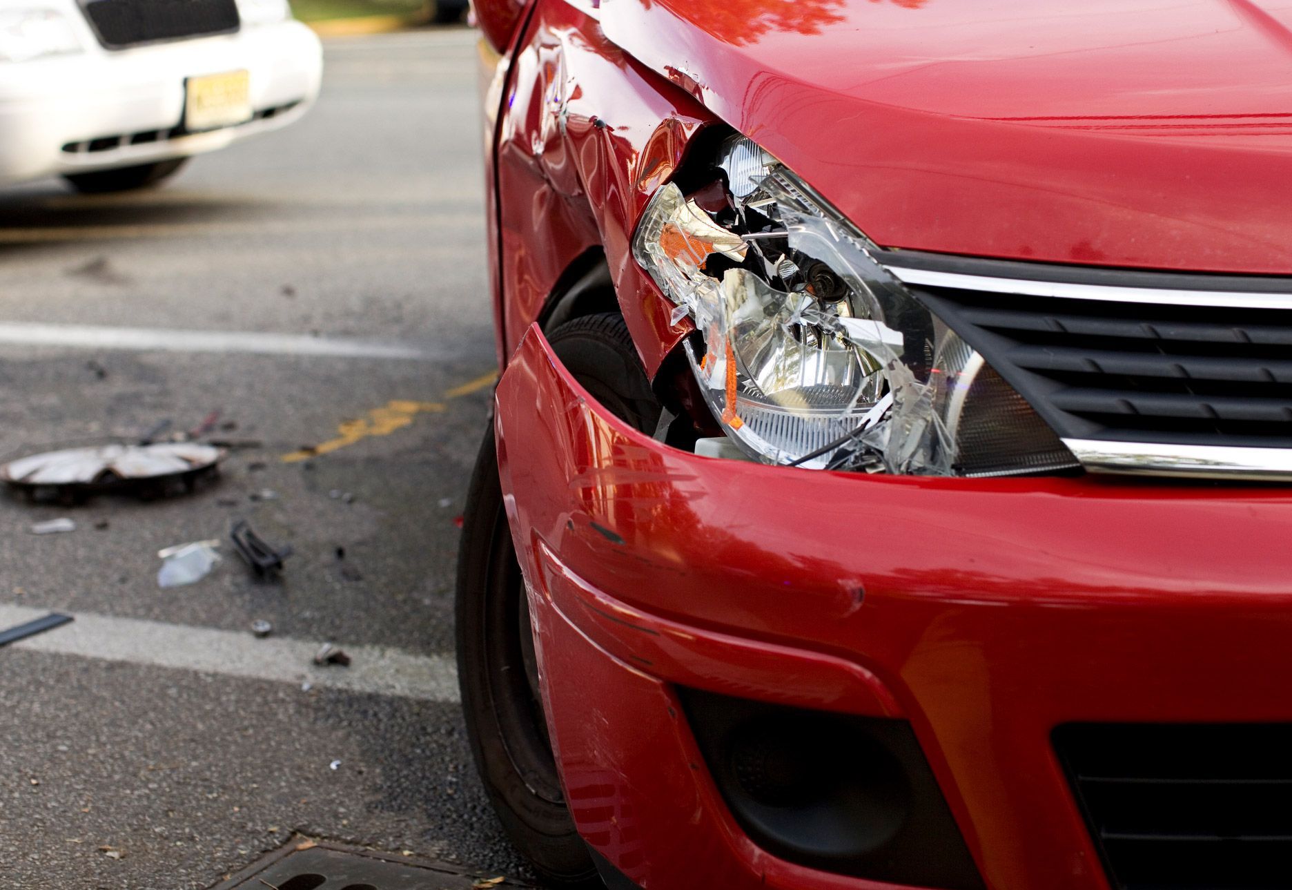 Red car with extensive front-end damage, broken headlight, parked on a street. A white vehicle is in the background.