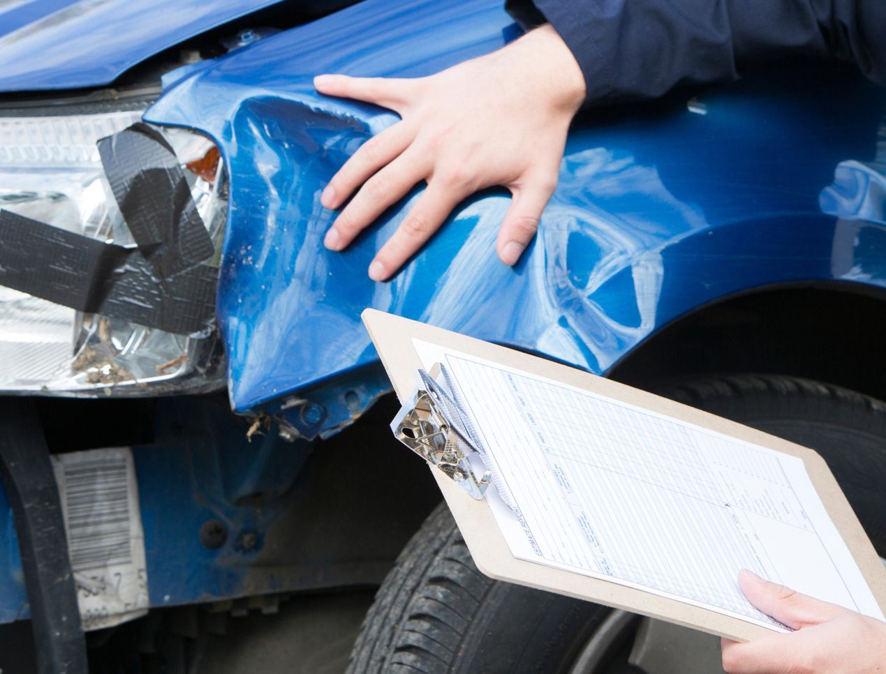 Person inspecting a damaged blue car's front with a clipboard, likely for an insurance claim.