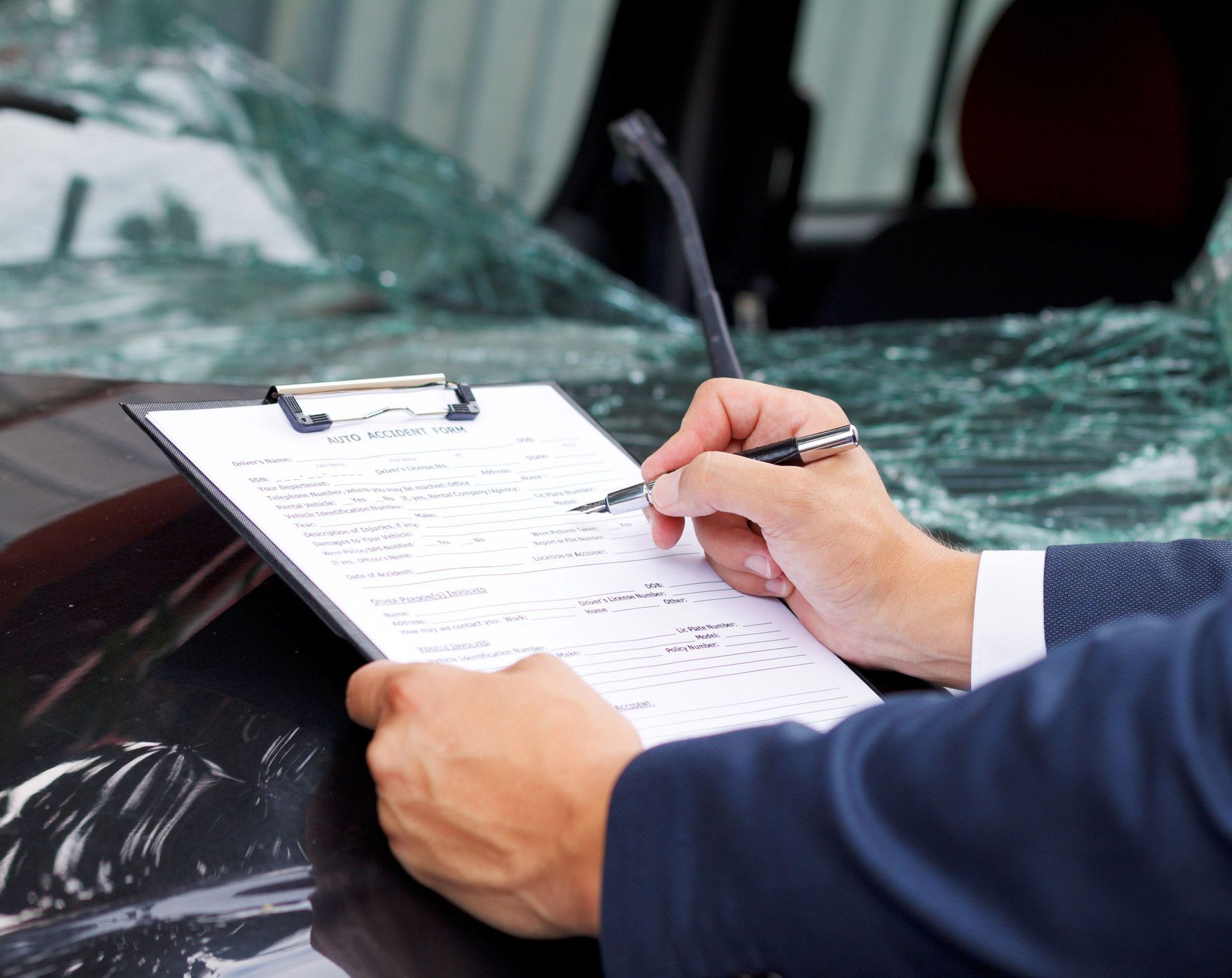 Person in suit writing on clipboard at a damaged car; broken windshield in the background.