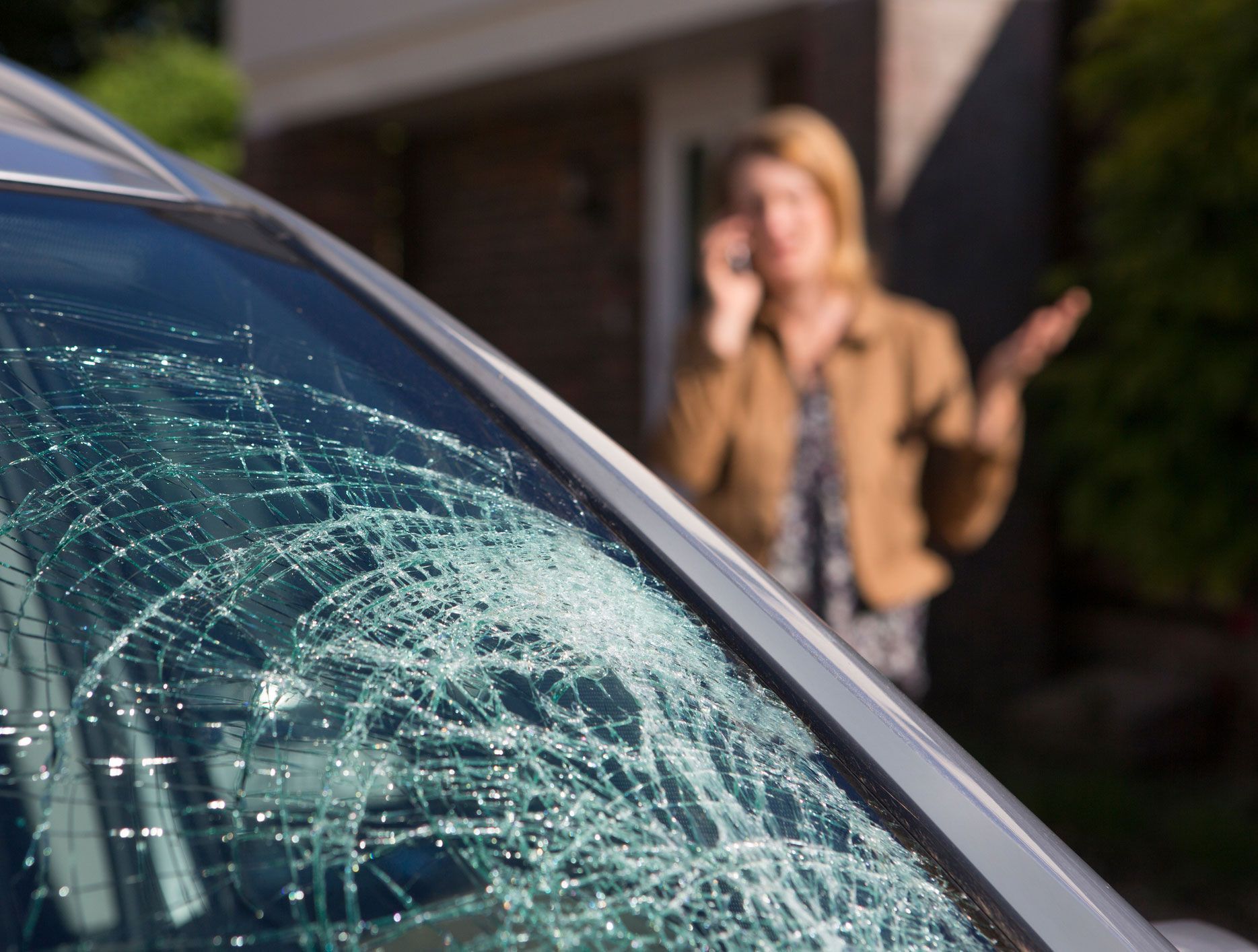 Shattered car windshield with a woman in the background, appearing distressed, talking on the phone outside a house.