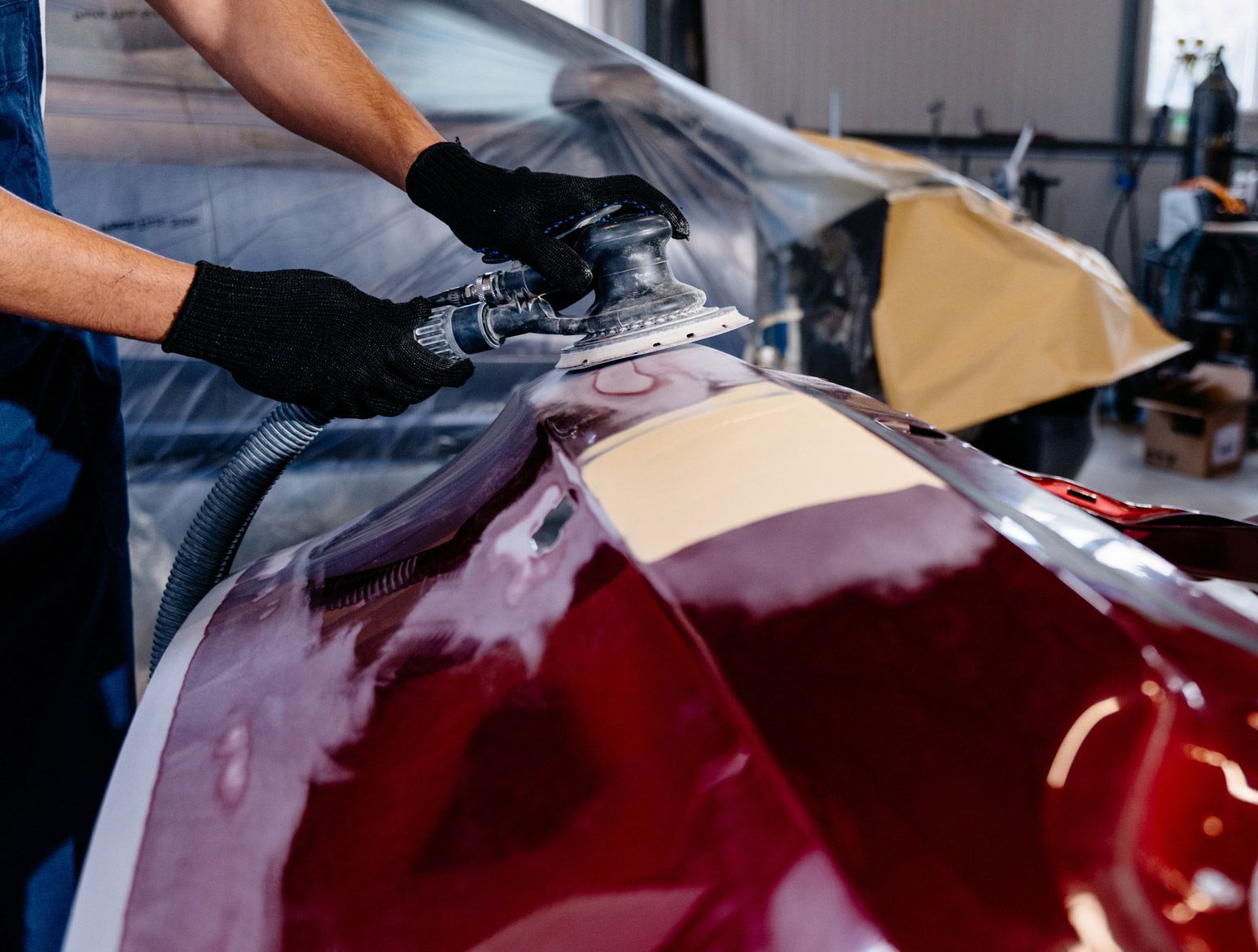 Person sanding a red car part with an air sander in a repair shop.