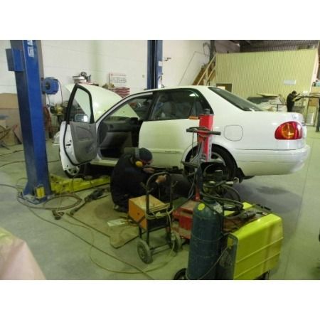 Mechanic working on a white car inside a garage; car is on a lift with a door open.