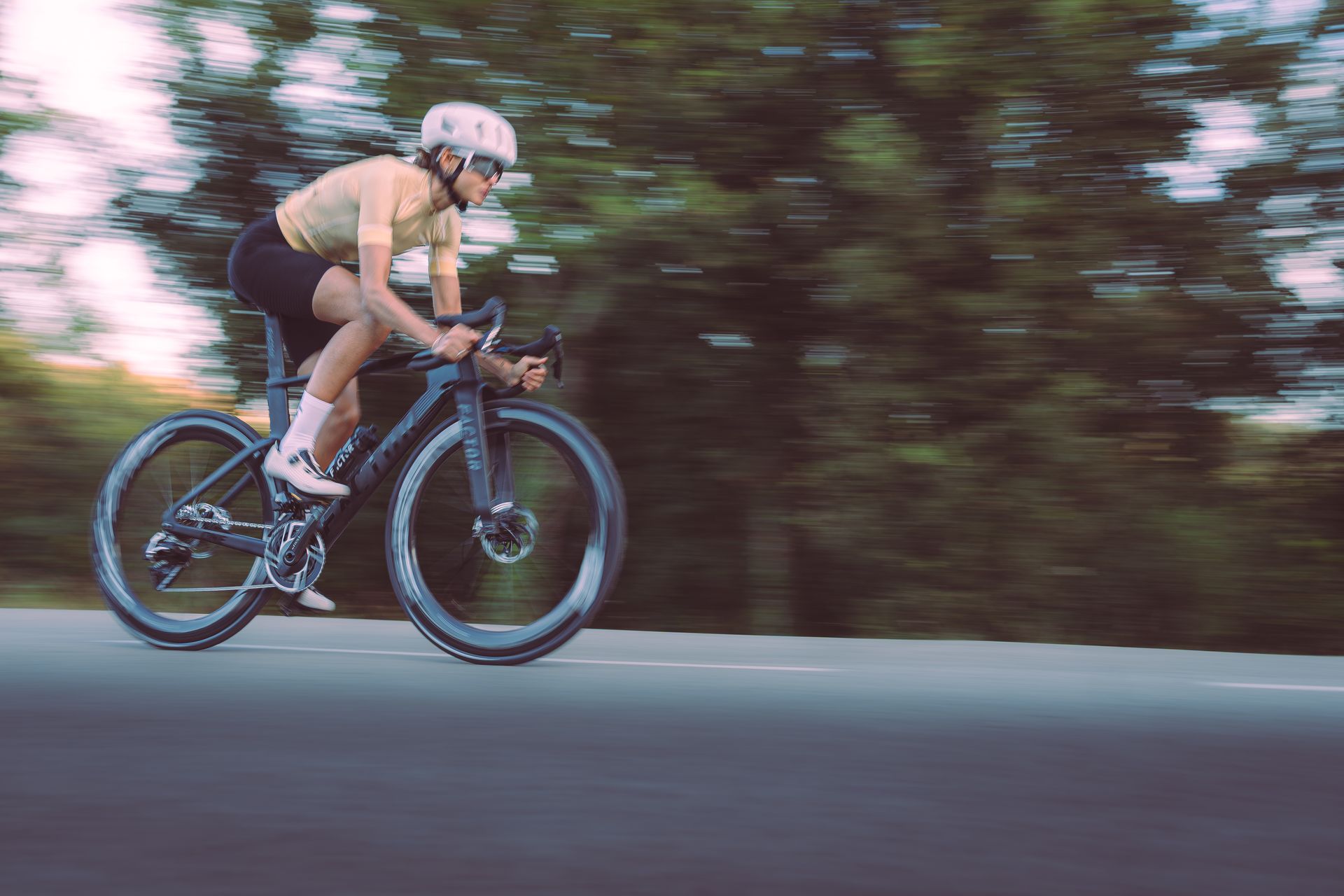 Cyclist in helmet and cycling gear rides a road bike on a paved road with blurred green trees in the background.