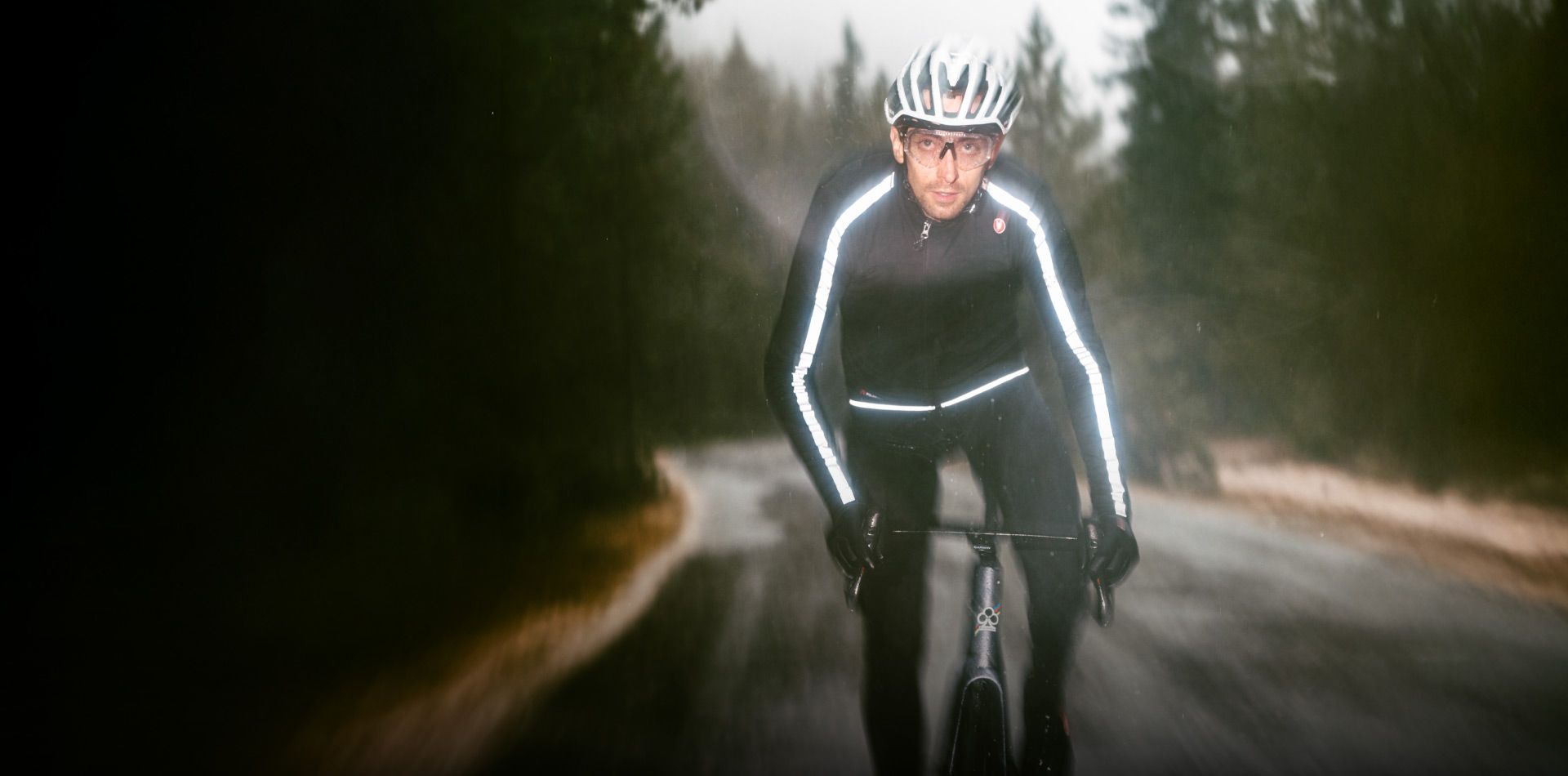 Cyclist riding on a wet road, wearing reflective black cycling gear and a helmet. Forest in background.