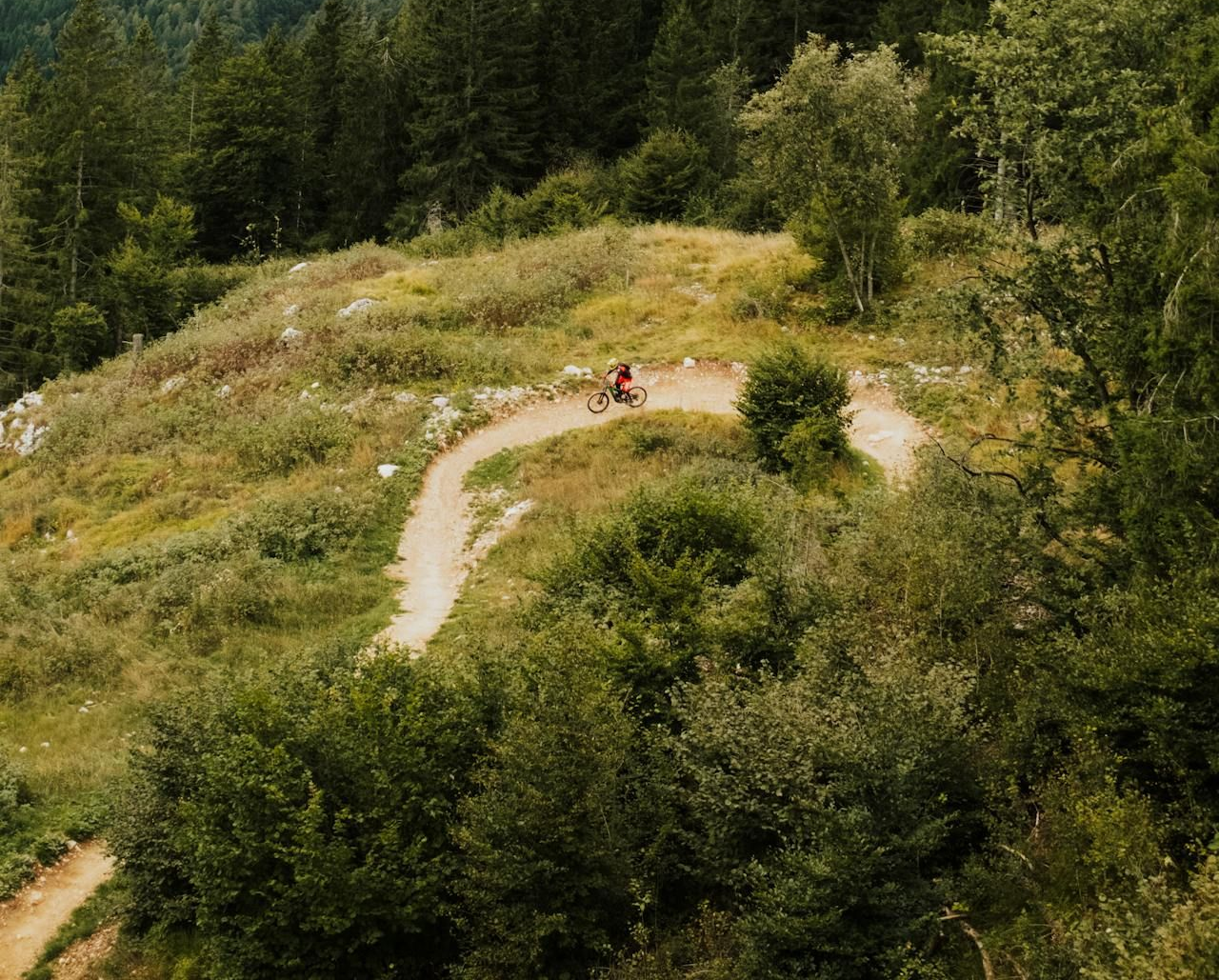 Dirt trail winds through green hillside, leading to a distant person cycling, with a rocky mountain backdrop.