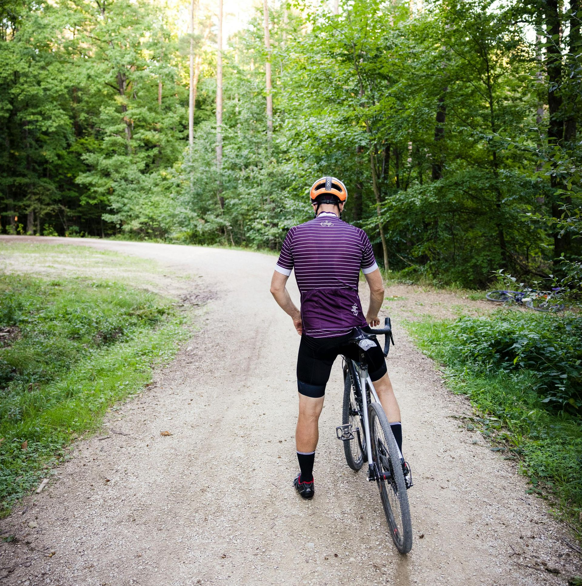 Cyclist standing on a gravel path in a forest, bike in hand, wearing a purple jersey and helmet.