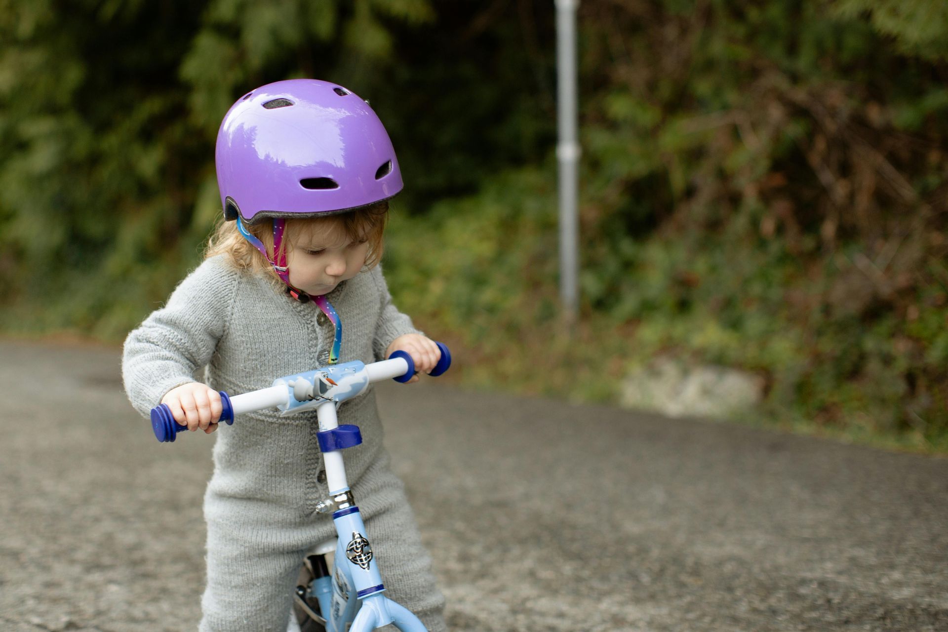 A toddler wearing a purple helmet and a gray jumpsuit holds the handlebars of a blue balance bike on a paved path.