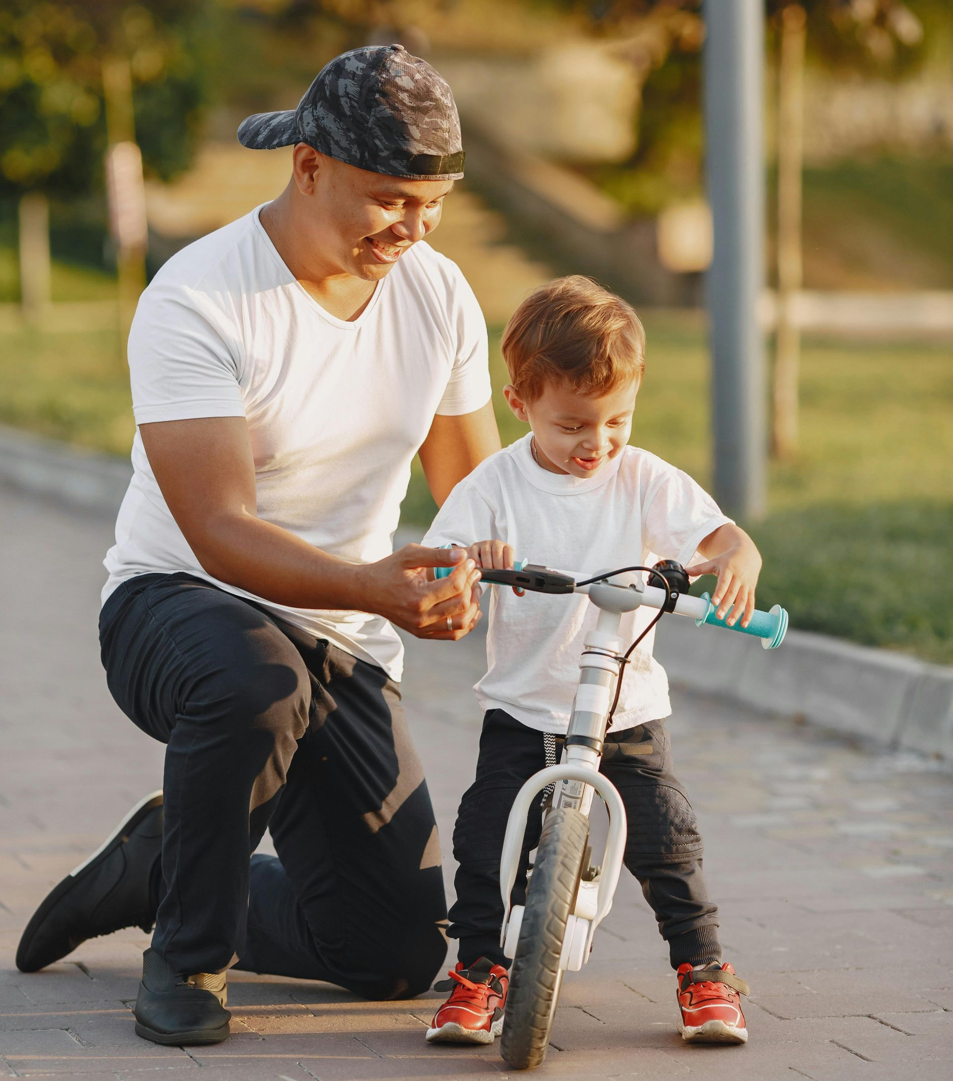 A man kneeling on a path to help a child balance on a bicycle outdoors.