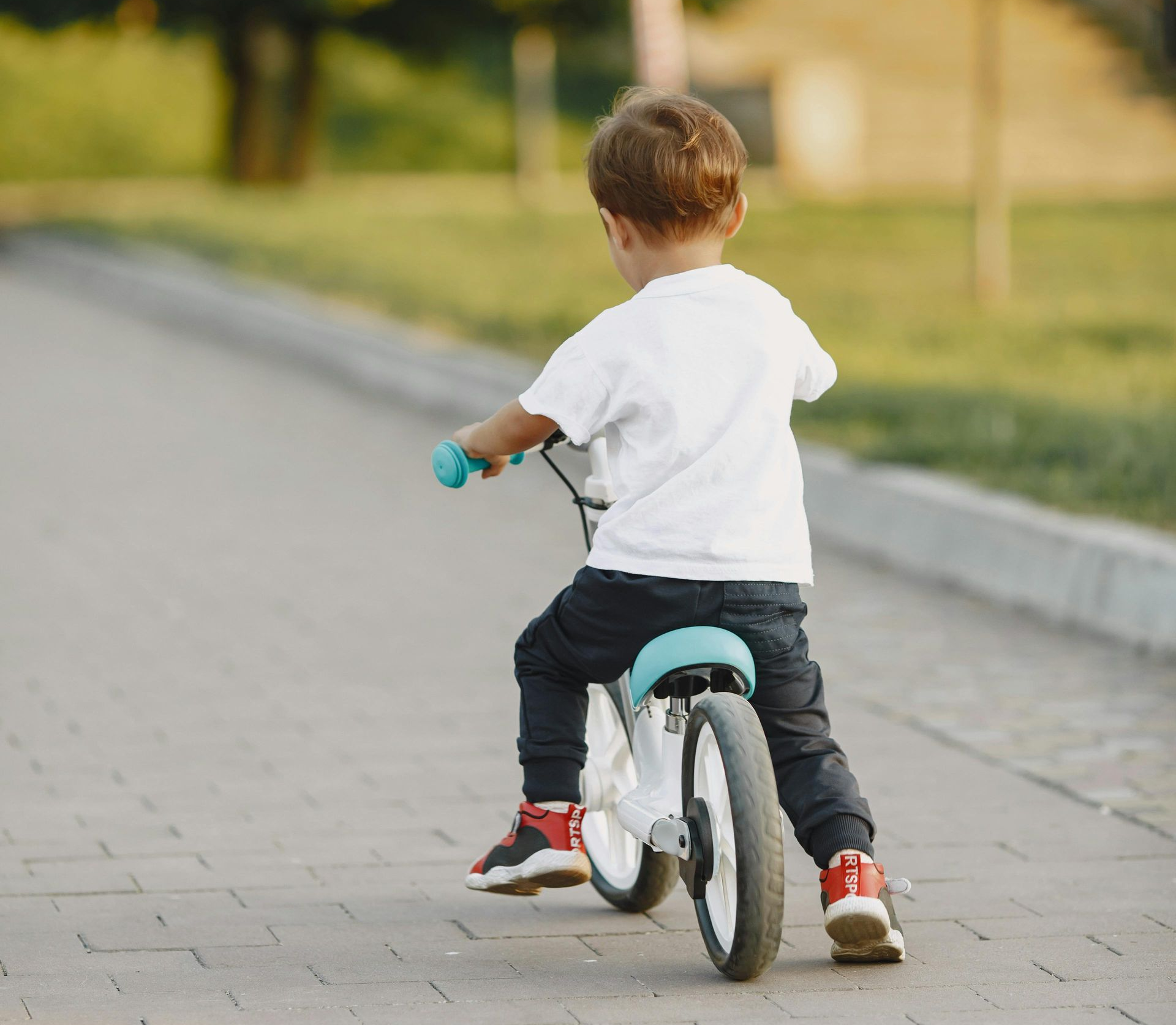 A child rides a balance bike down a paved path in a park during the late afternoon.