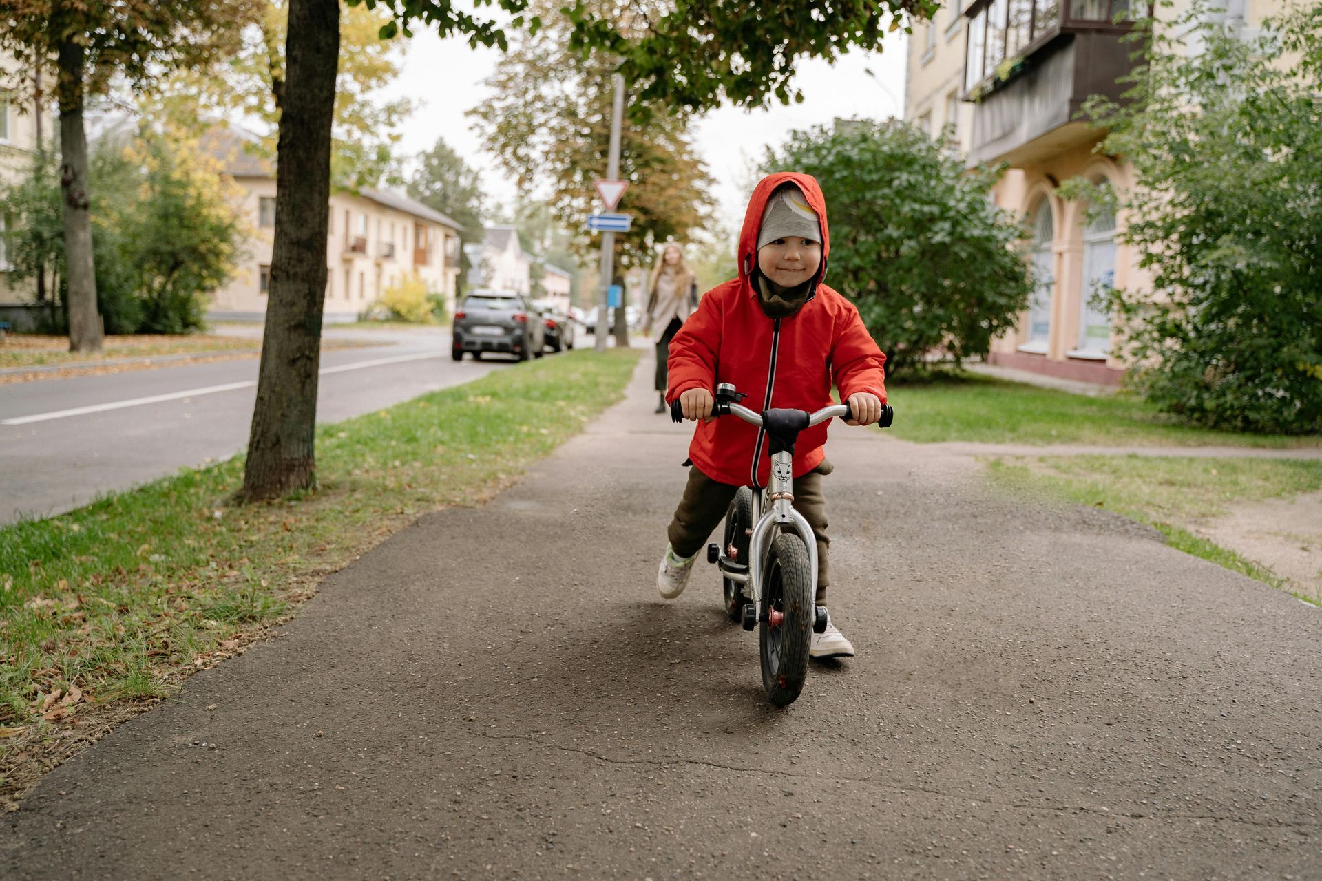 A toddler in a red hooded jacket rides a small balance bike on a paved sidewalk next to a street.