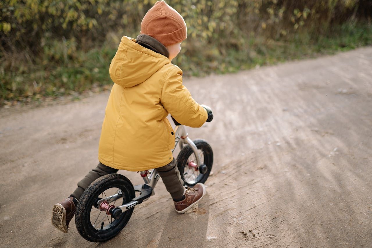 A person wearing a yellow jacket and brown beanie rides a balance bike on a dirt path.