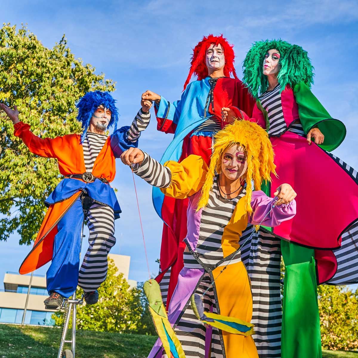 Clowns on stilts, colourful costumes and wigs. Outdoor park setting, blue sky.