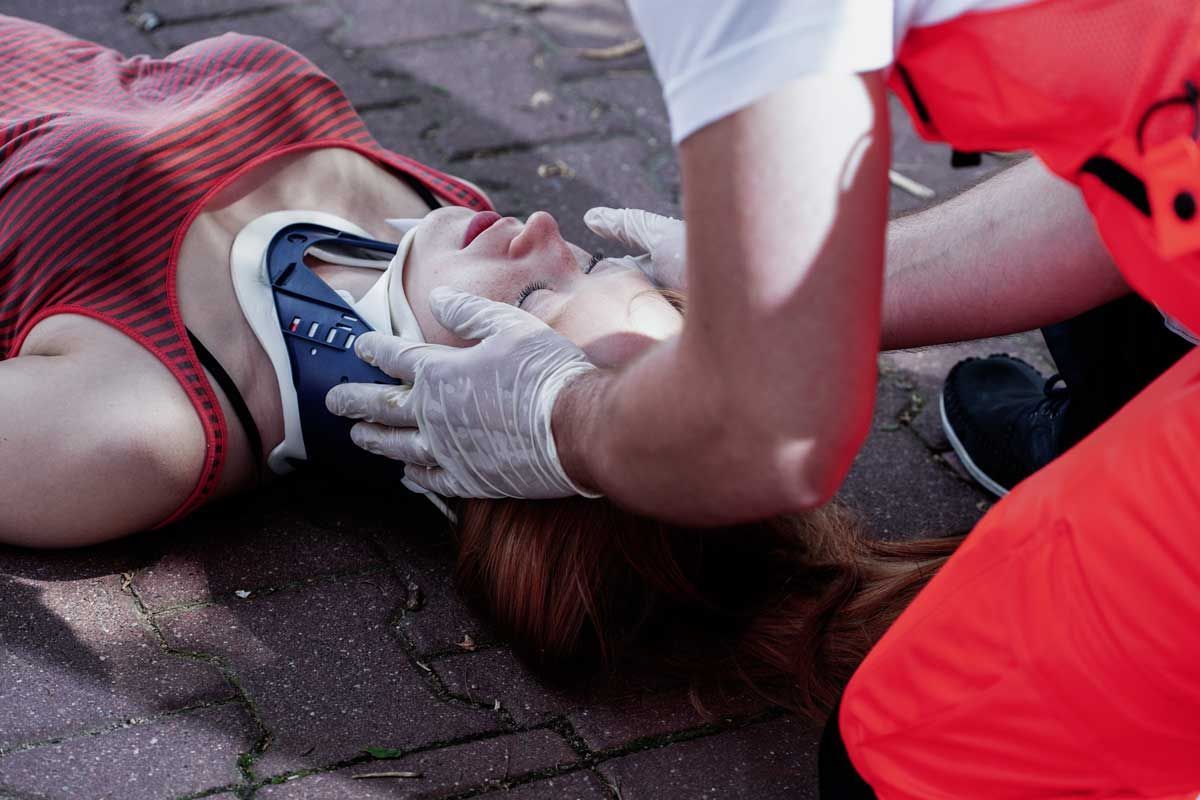 Paramedic attending to woman on the ground with neck brace. Red and orange uniform, grey gloves.