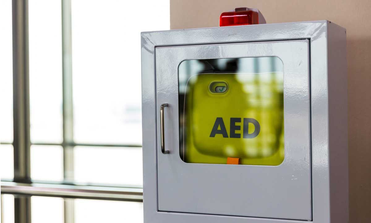 AED cabinet with a green defibrillator, red flashing light, and a handle.