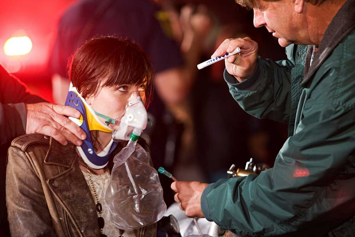 Woman with neck brace and oxygen mask, being assisted by a paramedic in an outdoor setting.
