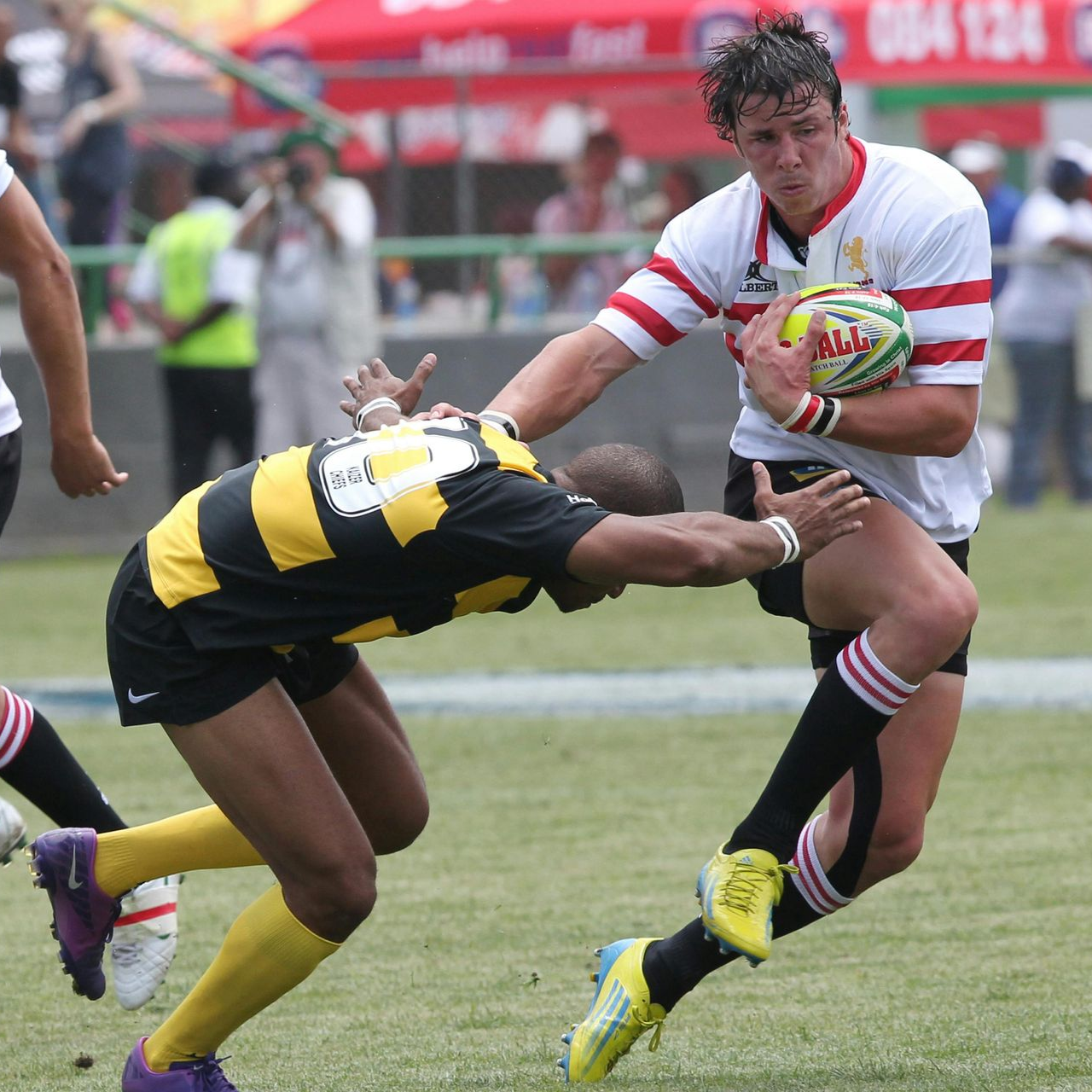 Rugby player in white jersey runs with ball, being tackled by a player in yellow and black jersey.