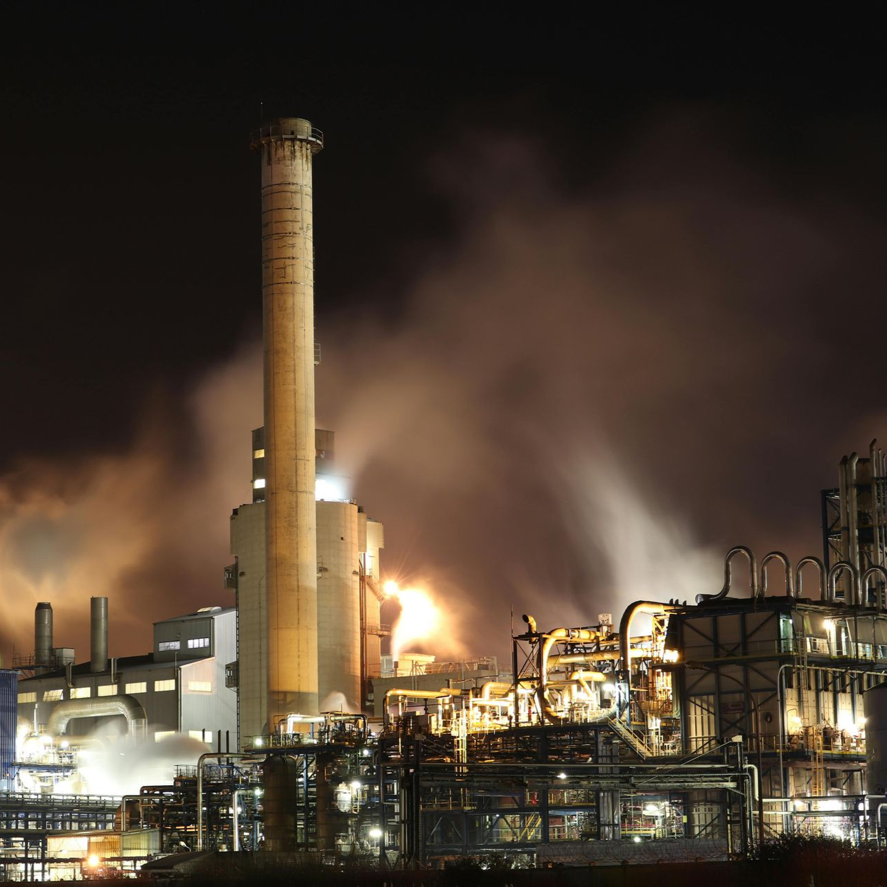 Factory at night with illuminated structures, emitting smoke and flames, tall smokestack.